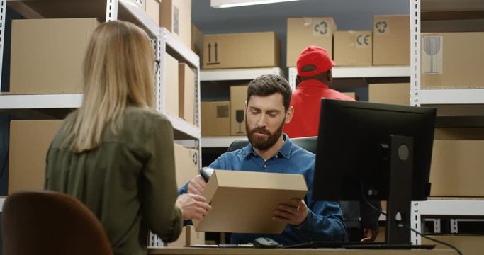 Caucasian handsome postman sitting at desk in postal store with boxes at computer and handing carton parcel to woman. African American mailman bringing box to female client of post office.