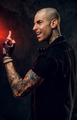 Bold and daring, tattooed, bald male model posing in a dark studio for the photoshoot wearing cool black denim vest with spikes, black shirt and leather gloves, rocker style, showing a gesture wirh