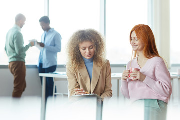 Portrait of two colorful young women discussing work project during coffee break in office, young intern presenting ideas to manager, copy space