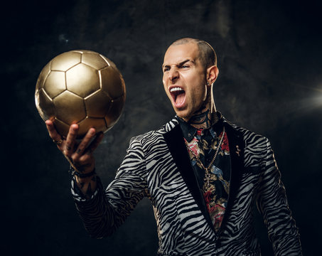 Expressive, fashionable, tattooed, bald male model posing in a studio for the photoshoot wearing fashionable custom made zebra style tuxedo, rose patterned shirt, looking on a golden soccer ball while