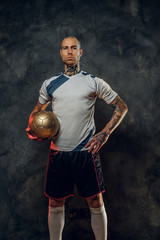 Bold and handsome soccer player posing for a photoshot in a dark studio, wearing professional sportswear, and holding a golden soccer ball, full height