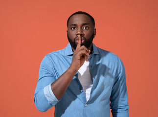 Man holds finger on lips and showing silence gesture. Photo of african man in casual outfit on coral background.
