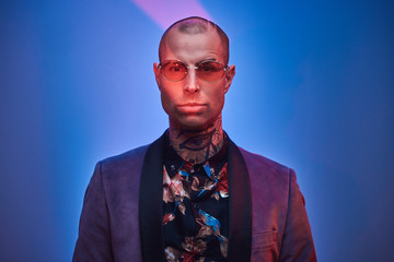 Obraz premium Cool, vivid, tattooed, bald male model posing in a studio for the photoshoot wearing fashionable custom made grey tuxedo, rose patterned shirt, and red glasses, vogue style