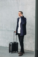 Traveler standing outdoors with suitcase and making call on cell phone