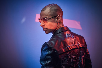 Cool, daring, tattooed, bald male rocker model posing in a studio for the photoshoot wearing fashionable designer black leather jacket, black shirt, red glasses, and jewelery, look from the back