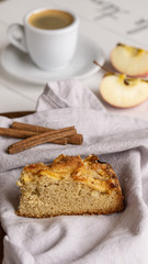 Apple pie slice lying on a grey cotton napkin. There is cinnamon sticks, apple and cup of coffee on the background. Wooden white background. 