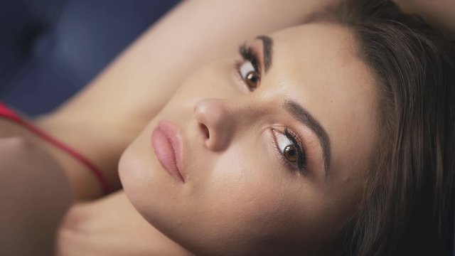 Beautiful young sexy woman attractive face gentle skin portrait curly hair brunette dressed in red silk dress on thin straps lying on the sofa gently stroking her hands over her shoulders and face.