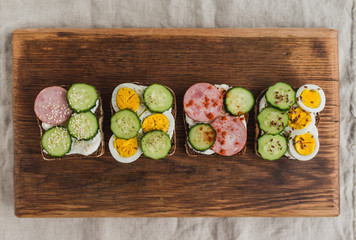 Sandwiches with rye bread, cream cheese, cucumber, egg, ham, spices and seeds. Set of delicious sandwiches on a rustic old board. Top view.