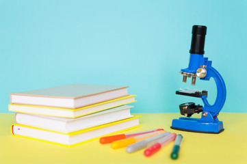 Blue microscope with felt-tip pens and books on a yellow table. The concept of schooling, research,...
