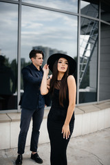 Obraz premium A beautiful, stylish pair of young people in black clothes and glasses stand against the background of an office building in the sunset. Fashion and style
