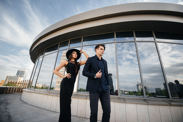 A beautiful, stylish pair of young people in black clothes and glasses stand against the background of an office building in the sunset. Fashion and style