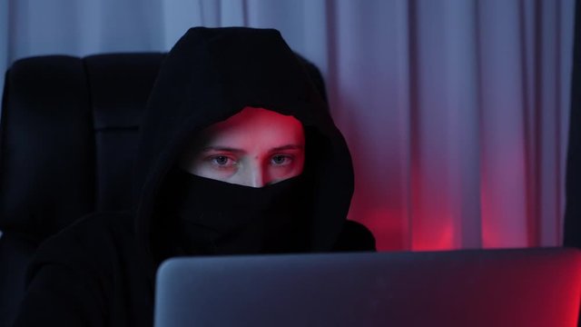 Woman face in black mask and hoodie looking at computer screen and breaking password. Young hacker breaks security protection on laptop screen. Concept of social security, online, virus protection