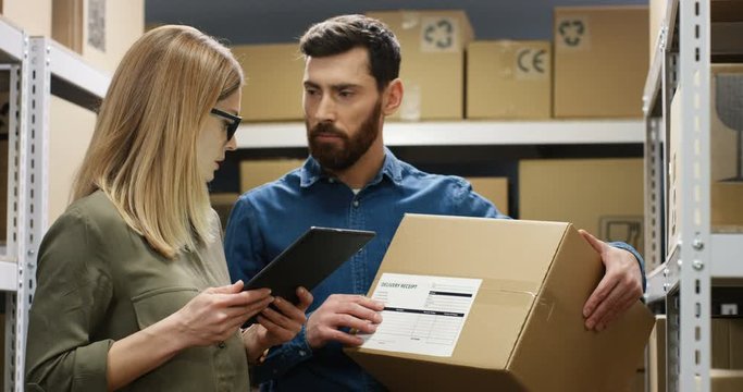 Caucasian young female post worker standing in postal store with parcels and tapping on tablet computer. Male co-worker courier coming with carton box and they registering parcel online on computer.