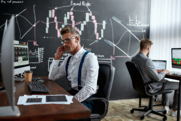 Invest Today, Earn Tomorrow. Stylish businessman, trader sitting in front of computer monitor while working using wireless earphones. His colleague and blackboard full of data analyses in background.