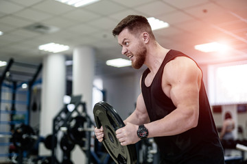 Naklejka na ściany i meble Portrait of handsome muscular powerlifter with weight disk in his hands in the sport gym. Bodybuilder with heavy disk trains his biceps with the open mouth. Close-up
