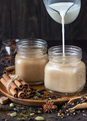 Indian masala chai, spicy tea with milk poured over and ingredients for making tea on a wooden plate, dark stone background.