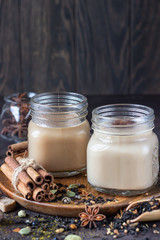Two mason jars with Indian masala chai tea on dark stone background. Spiced hot tea with milk and ingredients for making tea - cinnamon, anise, cardamom, nutmeg, pepper and brown sugar.