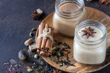 Two mason jars with Indian masala chai tea on dark stone background. Spiced hot tea with milk and ingredients for making tea - cinnamon, anise, cardamom, nutmeg, pepper and brown sugar.