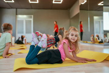 Kids and sport. Side view of a happy little girl smiling at camera while lying on yoga mat in the dance studio. Group of children doing stretching exercises with young female trainer