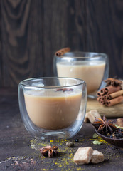Traditional masala chai in glass cup with ingredients (cinnamon, anise, cardamom, nutmeg, pepper and brown sugar) on a dark stone background. 