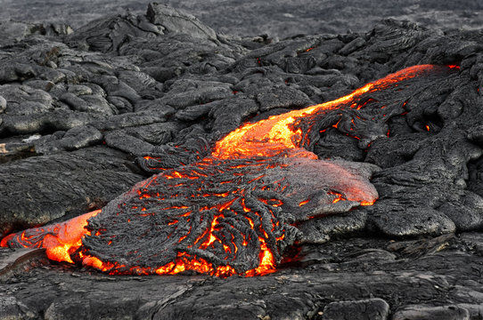Hot magma of an active lava flow emerges from a fissure and flows over previously deposited dark, strongly structured rock, the glowing lava shows itself in strong yellow and red tones