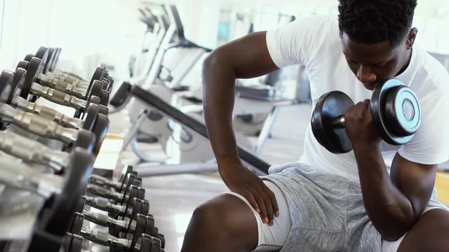 Young African American Man Sitting And Lifting A Dumbbell Close To The Rack At Gym. Male Weight Training Person Doing A Biceps Curl In Fitness Center.