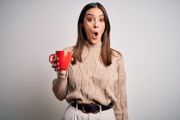 Young beautiful brunette woman drinking red cup of coffee over isolated white background scared in...