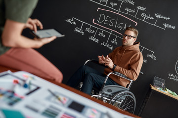 Creative people. Professional male UX UI designer in a wheelchair discussing new project with his colleague while sitting near blackboard with presentation in the modern office