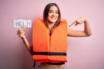 Young beautiful brunette woman wearing lifejacket holding paper with help word message with...