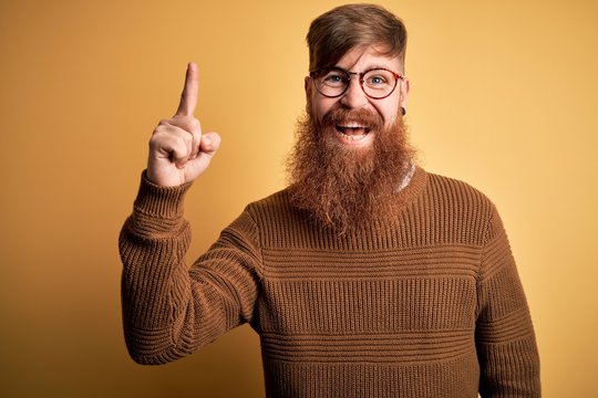 Handsome Irish redhead man with beard wearing glasses and winter sweater over yellow background pointing finger up with successful idea. Exited and happy. Number one.