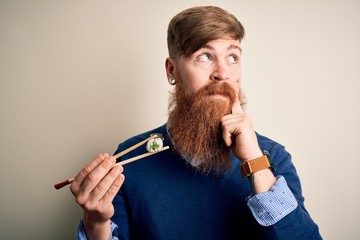Redhead Irish man with beard eating green maki sushi using chopsticks over yellow background...