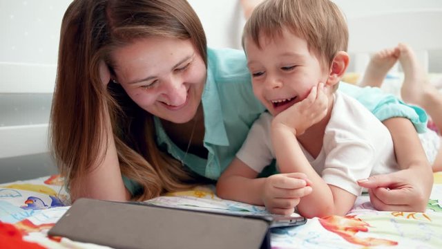 Closeup 4k footage of young mother with her little child son smiling and laughing while watching video on tablet computer before going to sleep
