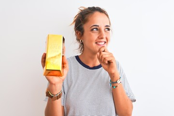 Young beautiful redhead woman holding gold ingot over isolated background serious face thinking about question, very confused idea