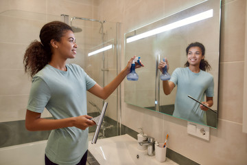 Making your home sparkling clean. Young smiling afro american woman in uniform cleaning a mirror with squeegee and spray detergent while working in the bathroom