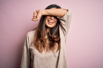 Young beautiful brunette woman wearing casual sweater standing over pink background covering eyes with arm smiling cheerful and funny. Blind concept.