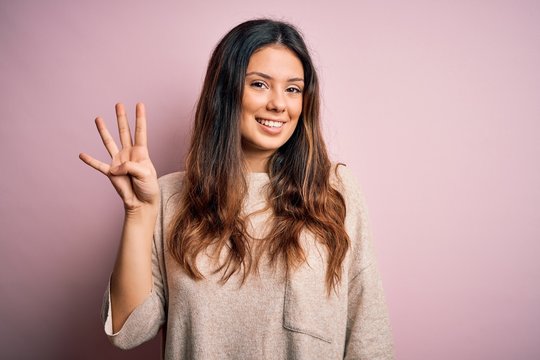 Young beautiful brunette woman wearing casual sweater standing over pink background showing and pointing up with fingers number four while smiling confident and happy.