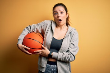 Young beautiful brunette sportswoman holding basketball ball over yellow background scared in shock...