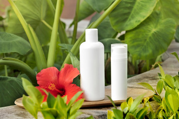 White empty tube of skincare bottle on bamboo plate with red hibiscus over tropical leaves background. Skincare spray tube on blue pool border.