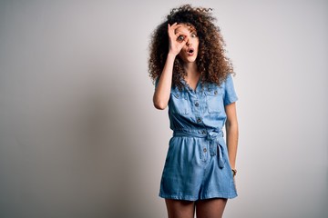 Young beautiful woman with curly hair and piercing wearing casual denim dress doing ok gesture shocked with surprised face, eye looking through fingers. Unbelieving expression.