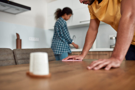 Clean home. Young couple in casual clothes cleaning kitchen together. Caucasian man cleaning kitchen table, while afro american woman washing dishes on the background - Powered by Adobe