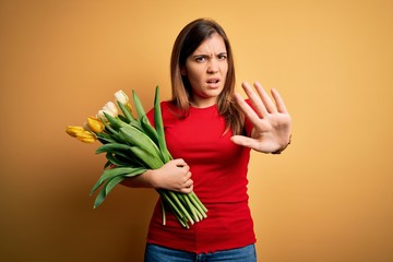 Young blonde woman holding romantic bouquet of tulips flowers over yellow background doing stop gesture with hands palms, angry and frustration expression