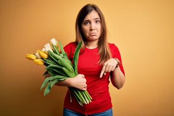 Young blonde woman holding romantic bouquet of tulips flowers over yellow background Pointing down looking sad and upset, indicating direction with fingers, unhappy and depressed.