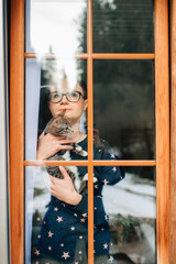 Handsome woman with pretty face holds a lovely cat on her hands and looks through the big window
