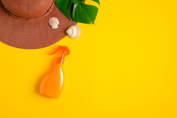 Sunscreen spray orange bottle, green tropical leaf, female beach hat on yellow background. Summer skin care cosmetic product and beach accessories. Top view with copy space