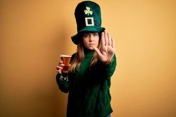 Young beautiful woman wearing green hat drinking glass of beer on saint patricks day doing stop...