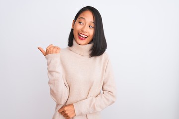 Young chinese woman wearing turtleneck sweater standing over isolated white background smiling with happy face looking and pointing to the side with thumb up.