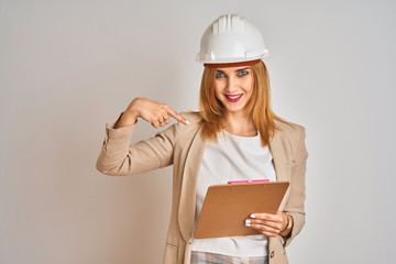 Redhead caucasian business woman wearing safety helmet and holding clipboard with surprise face pointing finger to himself