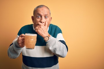 Senior handsome man drinking jar of beer standing over isolated yellow background shocked covering...