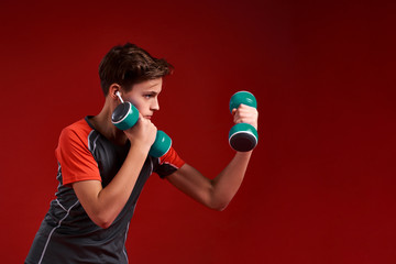 Motivation. A teenage boy engaged in sport, looking aside while exercising with dumbbells. Isolated on red background. Training, fitness, active lifestyle concept. Horizontal shot.
