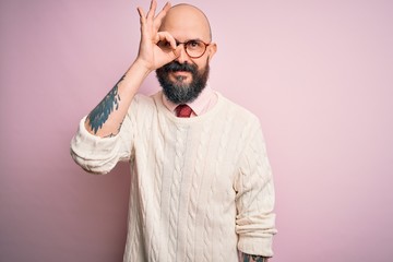 Handsome bald man with beard and tattoo wearing glasses and sweater over pink background doing ok gesture with hand smiling, eye looking through fingers with happy face.